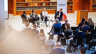 Vier Personen sitzen auf Stühlen vor einem Publikum in einem Raum mit Bücherregalen und einem Banner mit der Aufschrift 'COOPERATION DIALOGUE'
