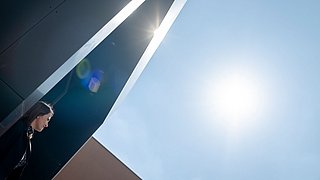 Eva Schmidt steht neben einem dunklen Gebäude, Sonnenstrahlen fallen durch eine Öffnung, blauer Himmel im Hintergrund.