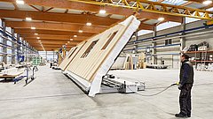 Person richtet maschinell ein vorgefertigtes Hausfassadenelement in einer Werkhalle auf.