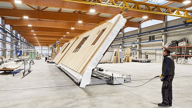 Person richtet maschinell ein vorgefertigtes Hausfassadenelement in einer Werkhalle auf.