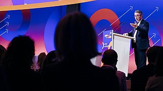 Mann im Anzug steht am Rednerpult vor Publikum, bunte Bühne mit Kreisen und Pfeilen im Hintergrund.