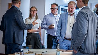 Mehrere Personen in Businesskleidung stehen um einen Tisch mit Getränken und Snacks und unterhalten sich in einem Büro.