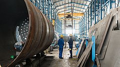 Zwei Arbeiter in blauen Overalls und gelben Helmen stehen in einer großen Fabrikhalle, umgeben von großen Metallrohren und Blechplatten.