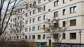 Mehrstöckiges Wohngebäude mit beige Fassade und vielen Fenstern, davor ein kleiner Gartenbereich mit Bäumen, Sträuchern, einer Pferdeskulptur und Gehwegen.
