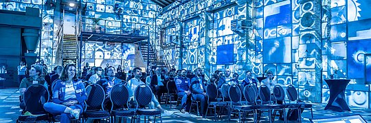 Menschen sitzen in einer Veranstaltungs- oder Konferenzhalle mit industriellem Flair, deren Wände und Boden durch blau-weiße Lichtprojektionen mit technischen Mustern und Symbolen beleuchtet sind.