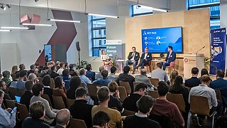 Vier Personen auf sitzen im Gespräch auf einer Bühne vor einem Publikum, im Hintergrund zeigt eine Präsentation Ansprechpartnerinnen des SET Hub