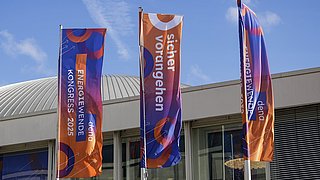Drei Fahnen mit orange-blauem Muster und den Texten 'ENERGIEWENDE KONGRESS 2025', 'sicher vorangehen' und 'dena' vor einem Gebäude mit gewölbtem Dach.