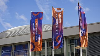 Drei Fahnen mit orange-blauem Muster und den Texten 'ENERGIEWENDE KONGRESS 2025', 'sicher vorangehen' und 'dena' vor einem Gebäude mit gewölbtem Dach.