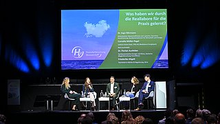 Vier Personene plus Moderatorin sitzen auf einem Panel auf der Bühne und diskutieren