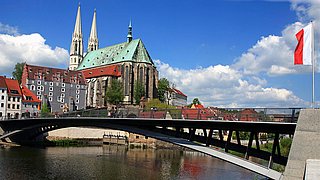 Blick auf die Stadt mit einer Brücke, einer Kirche mit grünem Dach und zwei Türmen, roten Dächern und einem Fluss im Vordergrund.