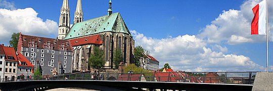 Blick auf die Stadt mit einer Brücke, einer Kirche mit grünem Dach und zwei Türmen, roten Dächern und einem Fluss im Vordergrund.