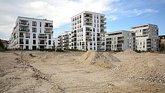 Moderne Wohngebäude mit mehreren Etagen, Balkonen und großen Fenstern, umgeben von unbebautem Gelände mit Erdhügeln.