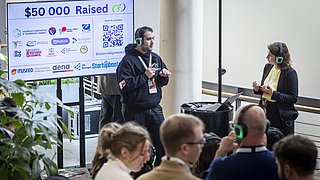 Mann mit Mikrofon und Frau mit Kopfhörern stehen vor Bildschirm mit Text '$50 000 Raised' und Logos, Publikum mit Kopfhörern im Vordergrund