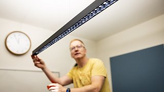 Eine Person hält eine lange, schwarze LED-Leuchte mit mehreren kleinen Lampen. Im Hintergrund ist eine Wand mit blauer Farbe und eine runde Wanduhr zu sehen.