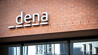 Logo von dena in moderner Schriftart, montiert an einer Ziegelwand. Darunter steht die Hausnummer '128 a'.