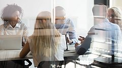 Eine Gruppe Büromitarbeiterinnen und Büromitarbeiter sitzen hinter einer Glasscheibe in einem Meetingraum für eine Besprechung.