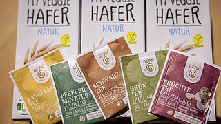 Drei Verpackungen mit der Aufschrift 'MY VEGGIE HAFER NATUR' und fünf Teebeutel mit den Bezeichnungen 'KAMILLEN TEE SANFT', 'PFEFFER MINZTEE WÜRZIG', 'SCHWARZ TEE KLASSISCH', 'GRÜN TEE MISCHUNG' und 'FRÜCHTE TEE MISCHUNG', alle in verschiedenen Farben.