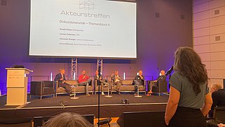 Sechs Personen sitzen auf einer Bühne in Sesseln und Sofas vor einer Leinwand mit der Überschrift 'Akteurstreffen', eine Frau steht mit Mikrofon im Publikum