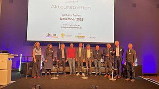 Gruppe von Menschen steht auf einer Bühne vor einer Leinwand mit der Aufschrift 'Akteurstreffen nächstes Treffen November 2026 Themenwünsche an info@akteurstreffen.de'