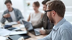 Mann mit Tasse im Vordergrund und drei Personen im Hintergrund an einem Tisch mit Laptop und Unterlagen