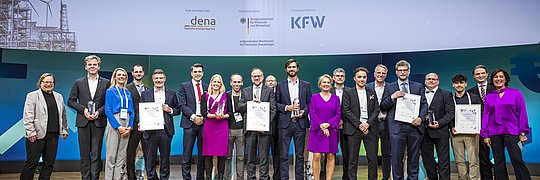 Gruppe von 17 Personen auf einer Bühne, die den 'Energy Efficiency Award 2024' halten. Im Hintergrund steht ein Banner mit den Logos von dena und KF