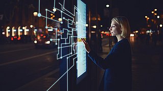 Eine Frau steht nachts vor einem beleuchteten interaktiven Touchscreen und berührt digitale Benutzeroberflächenelemente. Im Hintergrund sind eine Straße und Lichter einer Stadt zu sehen.