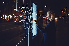 Eine Frau steht nachts vor einem beleuchteten interaktiven Touchscreen und berührt digitale Benutzeroberflächenelemente. Im Hintergrund sind eine Straße und Lichter einer Stadt zu sehen.