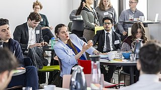 Mehrere Personen sitzen und stehen in einem modernen Konferenzraum, einige mit Notizblöcken und Getränken.