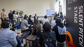 Mehrere Personen mit Kopfhörern sitzen und stehen vor einem Bildschirm auf der Silent Pitch Stage.