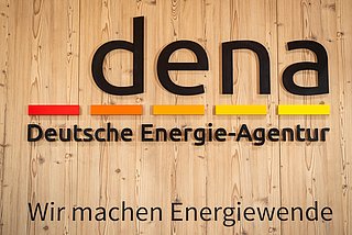 Logo der Deutschen Energie-Agentur (dena) in schwarzer Schrift auf Holzpaneel, darunter der Slogan 'Wir machen Energiewende'.