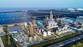 Industrieanlage mit rauchenden Kühltürmen, umgeben von großen Solarpanel-Feldern nahe der Küste.