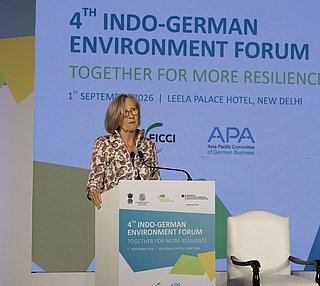 dena-Geschäftsführerin Kristina Haverkamp hält eine Präsentation auf der Bühne des 4. Deutsch-Indischen Umweltforums am 1. September 2026 in New Delhi. Im Hintergrund ist eine Leinwand mit dem Titel '4th Indo-German Environment Forum – Together for More Resilience' sowie Logos der veranstaltenden Organisationen zu sehen. Auf der Bühne stehen ein Rednerpult, Stühle und Mikrofone für eine Podiumsdiskussion."