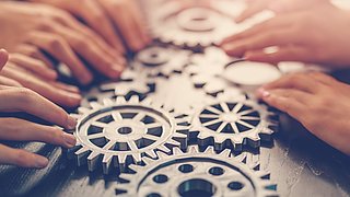 Mehrere Hände legen Zahnräder auf einen Holztisch, die ineinandergreifen.