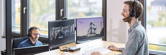 Person mit Headset auf dem Kopf steht an einem Schreibtisch mit zwei Monitoren während eines online Meetings.