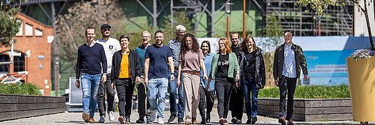 Gruppe von elf Personen unterschiedlichen Alters und Geschlechts, die auf einem gepflasterten Weg in einem urbanen Bereich mit Baustelle im Hintergrund gehen