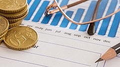 Stapeln von 50-Cent-Münzen neben einem Diagramm mit blauen Balken, einem Bleistift und einer Brille auf einem Blatt Papier.