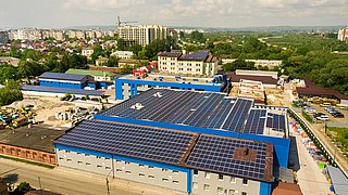 Luftaufnahme eines großen Gebäudes mit blauer Fassade und einem Dach, das mit Solarpanelen bedeckt ist. Im Hintergrund sind mehrere Wohngebäude und eine grüne Landschaft zu sehen.