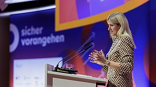 Frau mit langem blonden Haar und kariertem Blazer steht an einem Rednerpult mit zwei Mikrofonen vor einem bunten Hintergrund mit Texten.