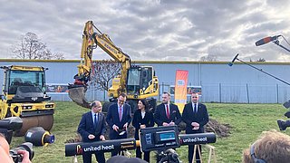 Fünf Menschen stehen um einen Tisch, dahinter stehen zwei gelbe Bagger