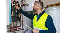 Mann mit gelber Warnweste stellt an Messgerät eines Heizungsrohrsystems ein und hält Tablet in der Hand.