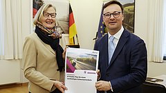 Kristina Haverkamp und Oliver Luksic in formeller Kleidung halten gemeinsam einen Bericht mit dem Titel '10-Punkte-Plan für einen nachhaltigen Schienenverkehr', im Hintergrund deutsche und europäische Flaggen