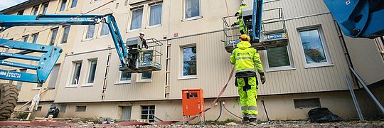 Montage eines vorgefertigten Fassadenelements an Mehrfamilienhaus, davor Kranfahrzeuge und Bauarbeiter
