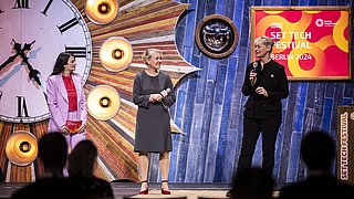 Das Bild zeigt drei Frauen auf einer Bühne, die am SET Tech Festival Berlin 2024 teilnehmen. Die Person in der Mitte ist die Vorsitzende der dena-Geschäftsführung Corinna Enders.