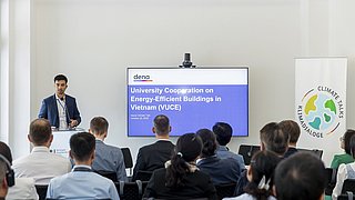 Tuan Ngo, Seniorexperte der Deutschen Energieagentur (dena) hält einen Vortrag vor Publikum in einem modernen Raum. Neben ihm steht ein großer Bildschirm, dort ist "University Cooperation on Energy-Efficient Buildings in Vietnam (VUCE)" zu lesen.