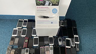 Eine Ansammlung von 40 Handys in verschiedenen Farben und Modellen, angeordnet auf dem Boden. In der Mitte steht ein Informationsschild mit dem Text: 'Die zweite Chance für Ihr Handy: Hier einwerfen, recyceln, wertvolle Rohstoffe gewinnen.'