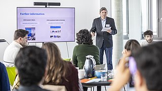 Ein Mann im Sakko hält einen Vortrag vor mehreren sitzenden Personen, während auf einem großen Bildschirm Präsentationsfolien mit dem Titel 'Further information' angezeigt werden.