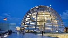 Die Reichstagskuppel in Berlin ist in der Abenddämmerung beleuchtet, in der Nähe weht eine deutsche Flagge und Besucher erkunden die Terrasse.