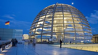 Die Reichstagskuppel in Berlin ist in der Abenddämmerung beleuchtet, in der Nähe weht eine deutsche Flagge und Besucher erkunden die Terrasse.