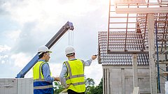 Zwei Bauarbeiter stehen auf einer Baustelle, umgeben von Baumaterialien, und besprechen die bevorstehenden Bauarbeiten.