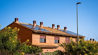 Reihenhäuser mit Solarmodulen auf Ziegeldächern, umgeben von Pflanzen und unter klarem blauem Himmel.
